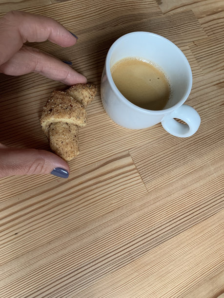 简约的木桌上放着一杯新鲜的浓缩咖啡，女性的手握着一个小可颂，完美适合温馨的早餐或咖啡馆相关的图片。图片下载