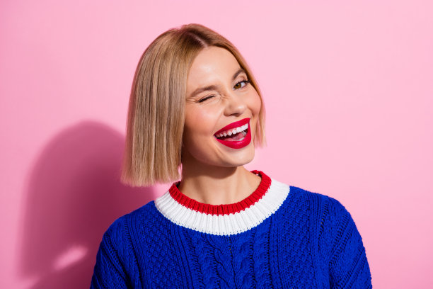 年轻迷人的金发女性模特在粉色背景上穿着蓝色针织毛衣，摆姿势并眨眼的肖像照片图片下载