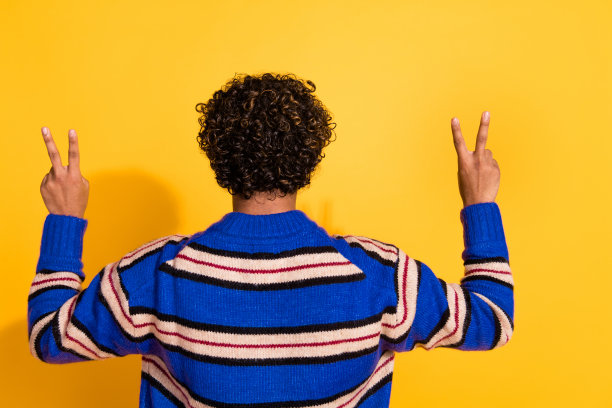 年轻帅气男性背影照片，做出V字手势，穿着时尚的蓝色条纹服装，背景为黄色。图片下载