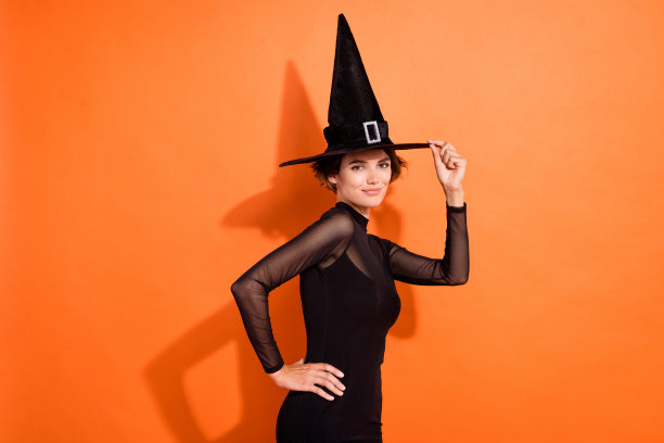 漂亮年轻女性的照片肖像，穿着时尚的黑色万圣节女巫服装，适合十月假期促销，背景为橙色。图片下载