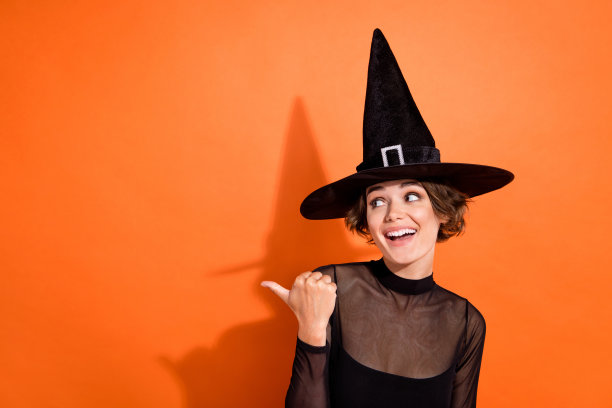 年轻女性的照片肖像，穿着时尚的黑色万圣节女巫服装，指向橙色背景上的空白空间。图片下载