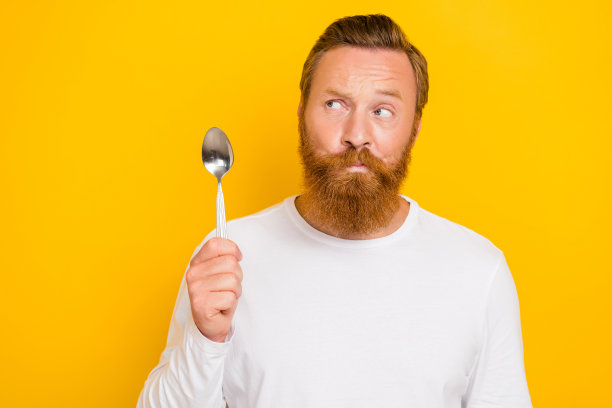 年轻男子的照片肖像，怀疑的表情，手持勺子，穿着时尚的白色服装，背景为黄色。图片下载