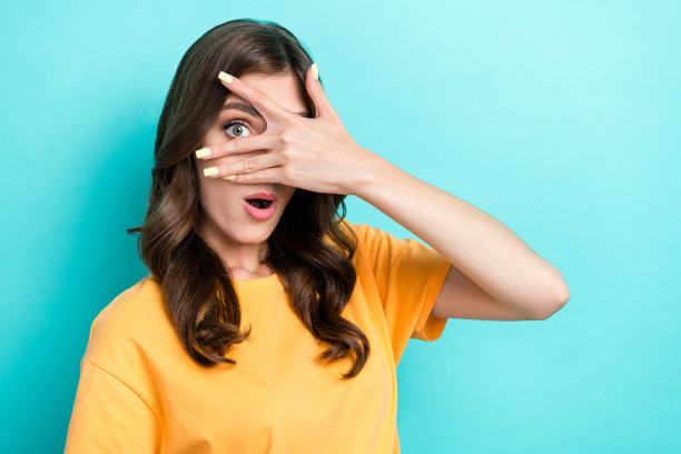 特写照片：年轻有趣的漂亮女孩穿着黄色T恤，遮住眼睛，张嘴露出兴奋、顽皮和震惊的反应，背景为水绿色。图片下载