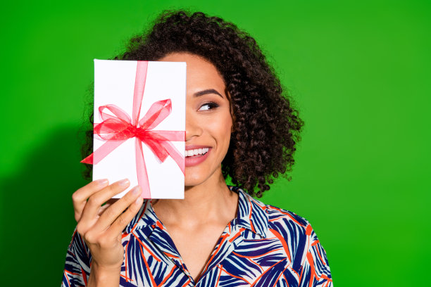 可爱的年轻女士的照片肖像，遮住脸部，手持礼物，目光投向空旷的空间，穿着时尚的彩色服装，背景为绿色。图片下载