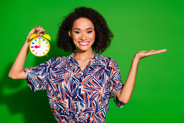 吸引人的年轻女性手持时钟，穿着时尚多彩的衣服，背景为绿色，留有空白空间的照片肖像。图片下载