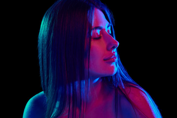 年轻女性的特写，的肩膀在黑色摄影棚背景下的明亮霓虹灯光中侧目。横向照片图片下载