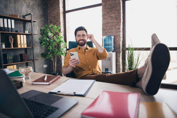 全身照片：一位年轻男性穿着黄色衬衫和正式服装，坐在桌子旁，手持设备，腿部交叠，展现出成功的商务人士形象，背景为明亮的办公室。图片下载