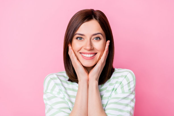 可爱年轻女孩的照片肖像，手掌触碰脸庞，享受完美柔软的肌肤，穿着时尚条纹服装，背景为粉色。图片下载