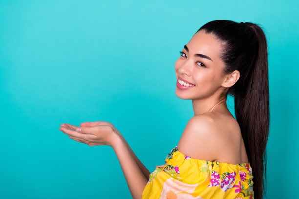 年轻漂亮的韩国女士近距离照片，手掌指向护肤产品，穿着时尚多彩的服装，背景为青色。图片下载