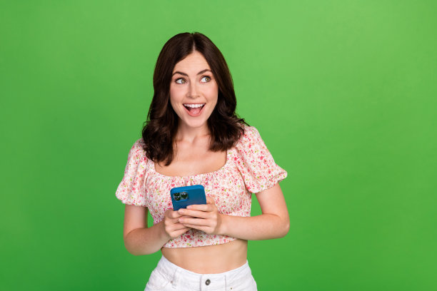 特写照片：年轻迷人、可爱的女士穿着粉色上衣，手持新智能手机，梦幻般地凝视着空旷的空间，适合广告促销，背景为绿色。图片下载