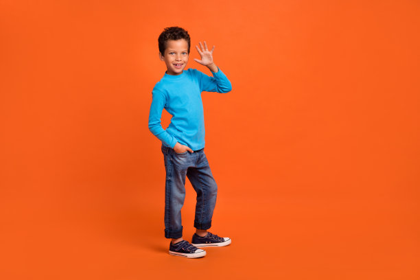 可爱的小男孩穿着时尚的蓝色服装，在橙色背景下挥手，展示购物横幅的全身照片图片下载