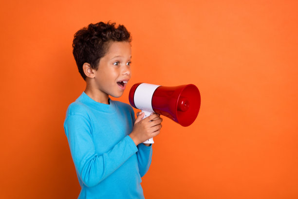 可爱的男孩在橙色背景下穿着时尚的蓝色衣服，手持扬声器进行宣布的照片肖像图片下载