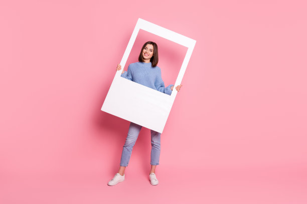 年轻漂亮的积极女孩在粉色背景下举着白色相框拍照的全身照片图片下载