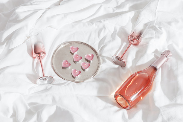 俯视闪亮的香槟杯、瓶装气泡玫瑰酒、白色床单上的粉色蜡烛心形装饰。极简生活美学照片，浪漫氛围，情人节，浪漫爱情概念。图片下载