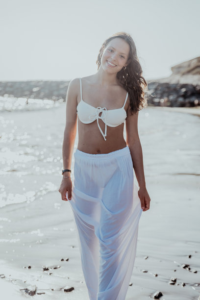 一位女性穿着白色比基尼和白色裤子在海滩上散步，竖版照片图片下载