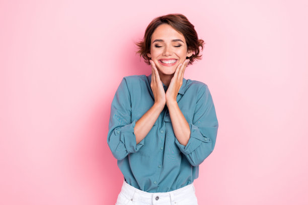 美丽年轻女性的照片肖像，闭眼触摸脸庞，享受柔软肌肤，穿着时尚的蓝色服装，背景为粉色。图片下载