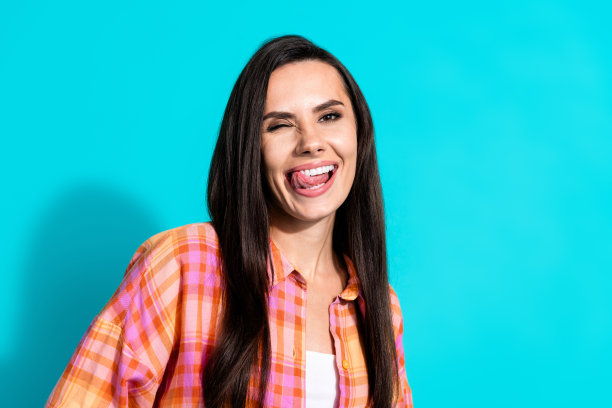 可爱的年轻女士在水绿色背景下，穿着时尚的格子服装，舔着牙齿并眨眼的照片肖像。图片下载