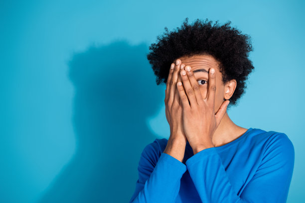 吸引人的年轻男子在蓝色背景下，穿着时尚衣服，遮住脸部，偷偷窥视的照片肖像。图片下载