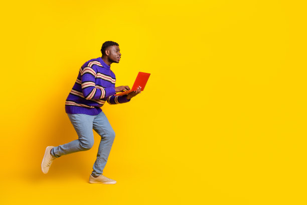 年轻男子在黄色背景下空旷的地方奔跑，手中拿着笔记本电脑的全身照片图片下载
