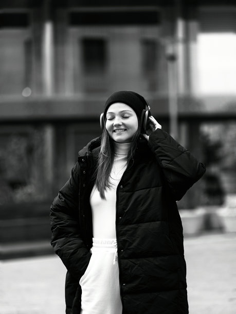 女孩戴着大耳机听音乐。微笑的女孩在户外放松，手里拿着智能手机和耳机。时尚女孩的肖像，背景是现代街道建筑。黑白竖版照片。图片下载