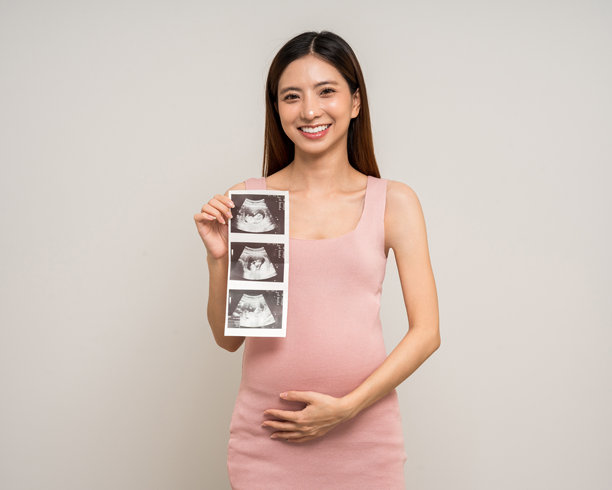 快乐的亚洲孕妇轻抚肚子，手中拿着超声波照片，背景为单色。家庭计划、母婴保险与孩子健康的孕期生活。图片下载