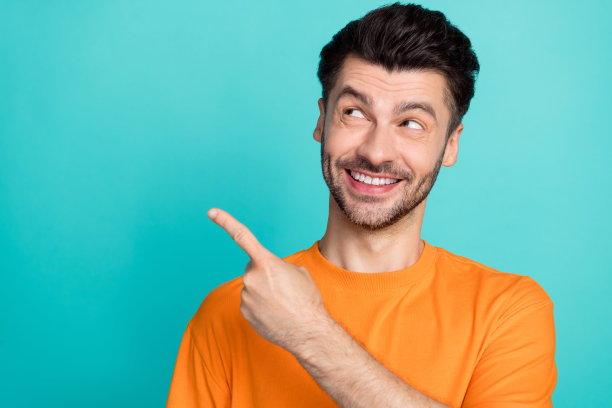 特写照片：年轻帅气、幽默、兴奋的男人穿着橙色T恤，手指指向空白处，表现出感兴趣的样子，背景为青色。图片下载