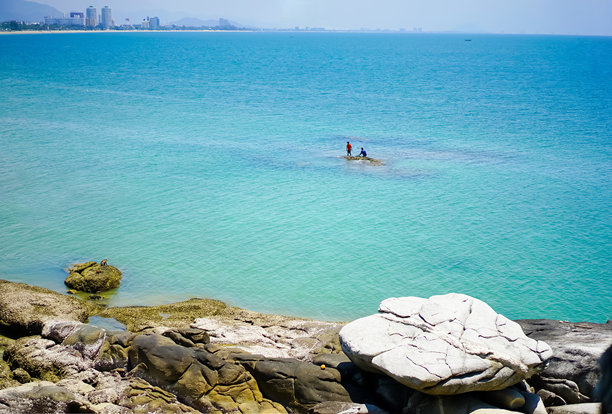 泰国华欣宁静海滩和蔚蓝海洋的风景照片，地平线上有摩天大楼的景观和蓝天。沙滩上的岩石和热带植物。图片下载