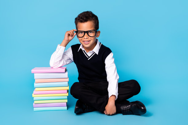 穿着黑色马甲和眼镜的可爱小男孩在蓝色背景下阅读书籍，心情愉快的全身照片图片下载
