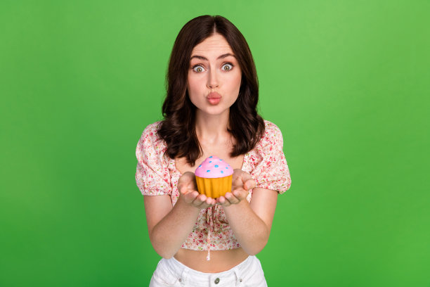 特写照片：年轻漂亮的女性撅嘴，手里拿着美味的松饼和蛋糕，做出空吻的姿势，时尚的女友，背景为绿色。图片下载