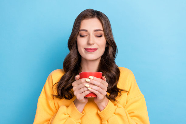 美丽年轻女孩的照片肖像，闭着眼睛享受咖啡香气，穿着时尚的黄色服装，背景为蓝色。图片下载