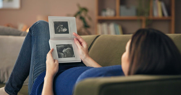 孕妇在沙发上放松，手持超声波图，思考和进行产前护理。未来的妈妈，宝宝的照片，正在客厅休息，为母爱、试管婴儿生育或成长准备。图片下载