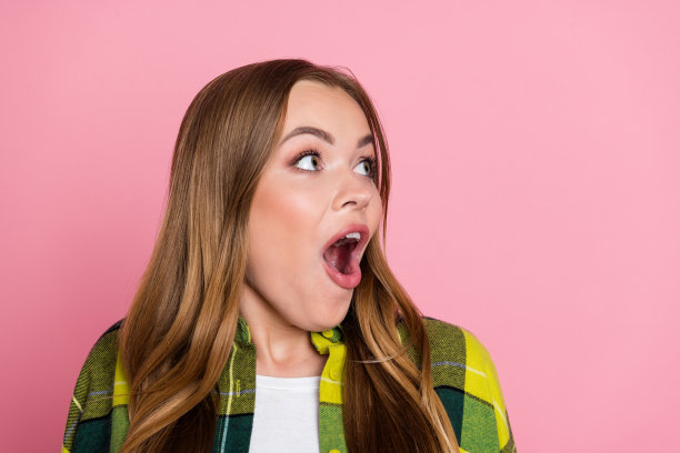 穿着黄色格子衣服的漂亮年轻女孩在粉色背景前惊讶地看着空旷的空间的照片肖像图片下载