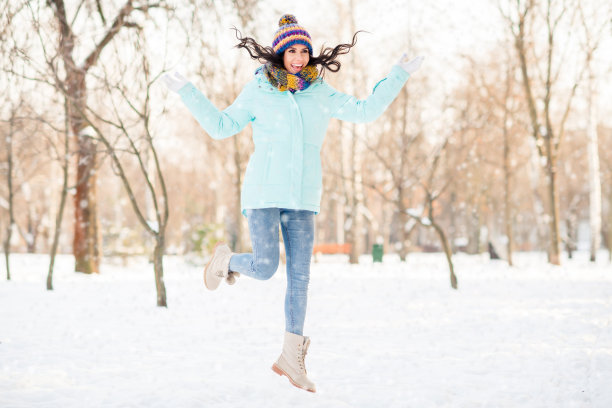 冬季公园户外，运动风格可爱的女士穿着针织手套、头饰、围巾、外套、牛仔裤和靴子跳跃的照片图片下载