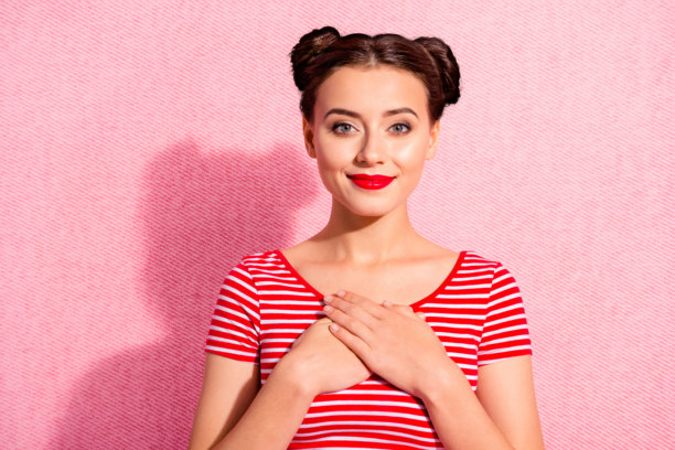 特写照片，漂亮的女士，迷人的发型，两个发髻，红色唇膏，双手交握，手臂放在心口，闭着眼睛，满脸喜悦，穿着休闲的红白条纹T恤，背景为粉色。图片下载