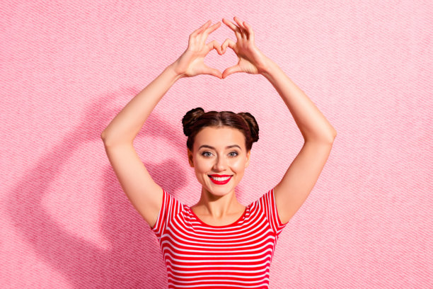 特写照片：美丽的女士，两个发髻，涂着唇膏，双手交叉成心形，手臂在头顶上方，展现心脏健康，穿着休闲的红白条纹T恤，背景为粉色。图片下载