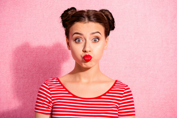 特写照片：美丽的女士，俏皮的发型，两个发髻，明亮的发蜡，诱人的大嘴唇，像孩子一样玩耍，穿着休闲的红白条纹T恤，背景为粉色。图片下载