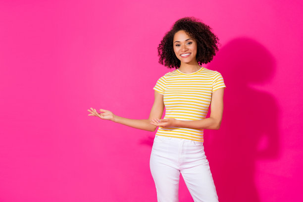 迷人的年轻女孩的照片肖像，手掌指向空旷的产品展示空间，穿着时尚的条纹服装，背景为明亮的粉色。图片下载