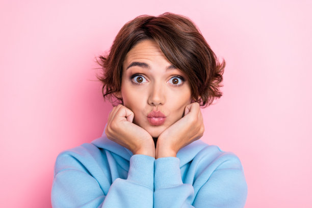 特写照片：年轻美丽的女孩穿着蓝色毛衣，双手托着脸颊，撅嘴送空气之吻，调皮可爱，背景为粉色。图片下载