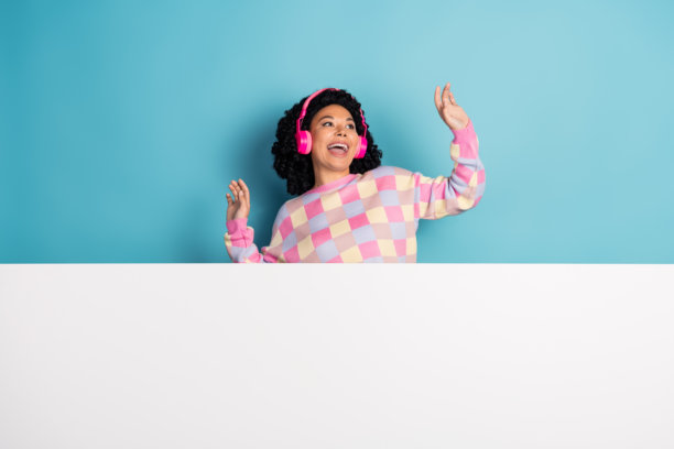 漂亮年轻女孩戴着耳机在蓝色背景下跳舞，穿着时尚的粉色格子服装，白色空旷空间中的照片肖像。图片下载