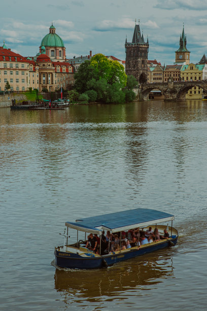 这是查尔斯桥和老城桥塔的美丽全景，背景是伏尔塔瓦河，前景有一只小蓝色渡船。照片拍摄于一个晴朗的日子，蓝天上飘着几朵云，增添了天空的层次感。图片下载