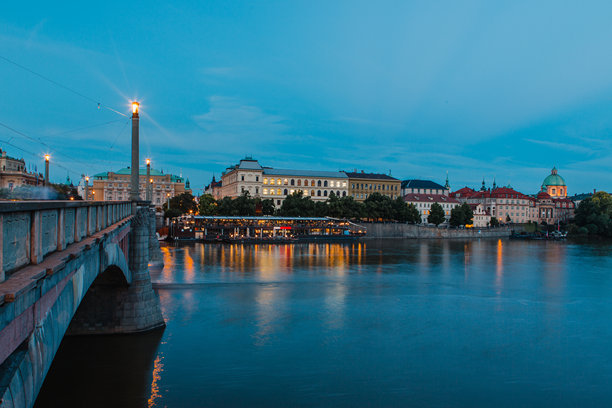 黄昏时分（落日后）伏尔塔瓦河上的桥梁照片。图像中深蓝色的夜空倒映在水面上，桥梁和城市的历史建筑在夜灯下熠熠生辉。图片下载
