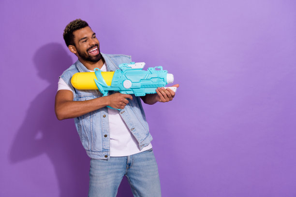 穿着牛仔背心的甜美男孩在紫色背景下空旷的地方用水枪拍照，显得很惊讶。图片下载