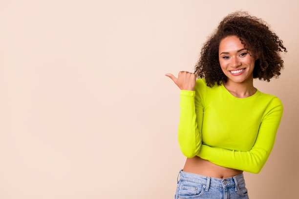 年轻可爱的女孩穿着黄色上衣，指向空白处，露出洁白的牙齿，特写照片，背景为米色。图片下载