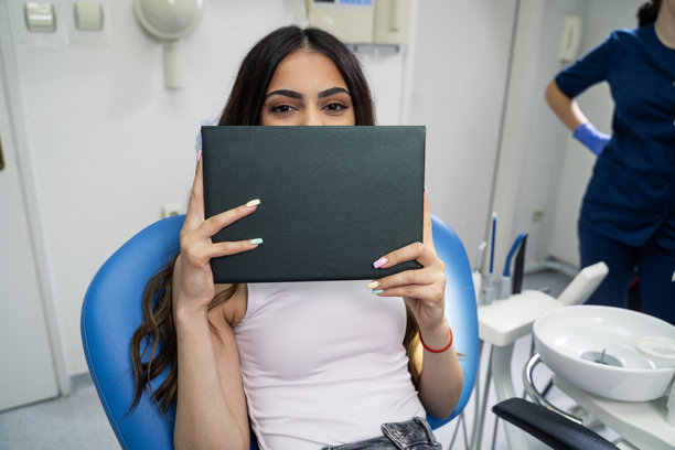 年轻女性坐在牙科椅上，手里拿着深绿色的文件夹遮住脸部，留有空白空间可添加文字或图片。图片下载