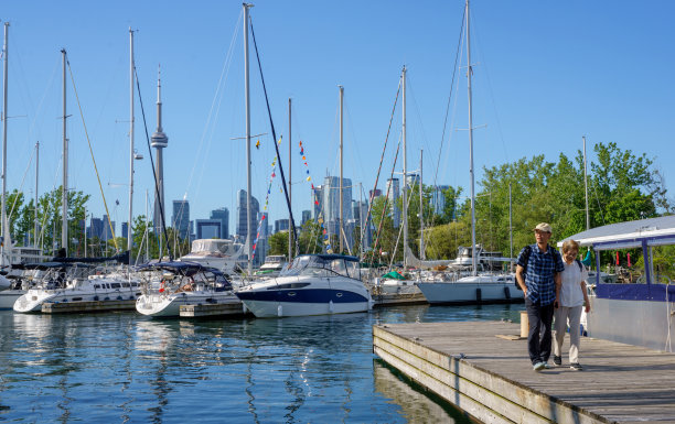 一对老年游客夫妇在多伦多中心岛上用手机拍照，背景是加拿大国家电视塔和多伦多市中心。图片下载
