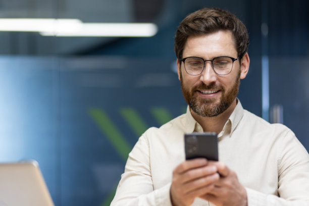 年轻商人在办公室的工作场所里，戴着眼镜和胡须，正坐着使用手机的特写照片图片下载