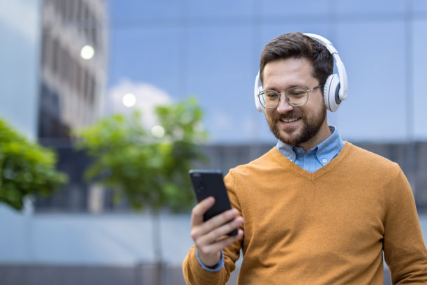 年轻男子在街上靠近办公室，戴着白色耳机，微笑着通过手机进行视频通话的特写照片图片下载
