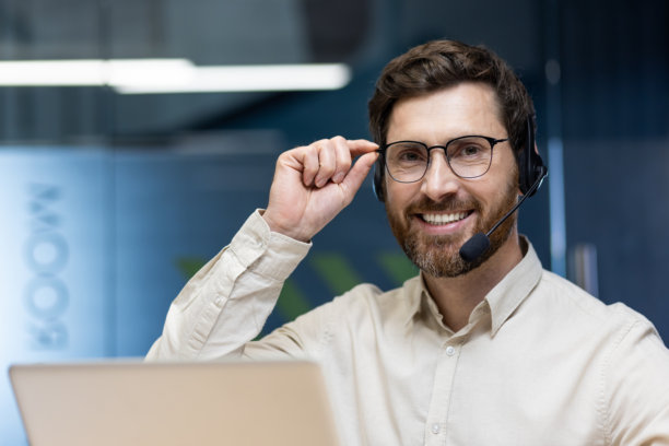 年轻男子戴着耳机坐在前台办公室的桌子旁，微笑着看向镜头，同时手里拿着眼镜，特写照片。图片下载