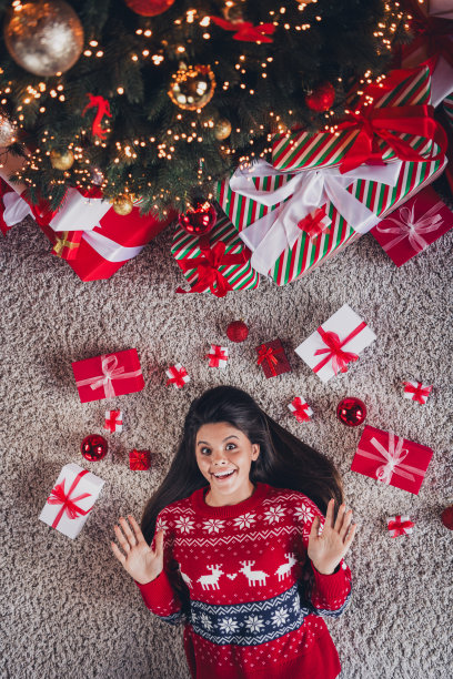 俯视照片中，快乐可爱的女孩穿着新款红色印花衣服，躺在地板上享受室内的圣诞节装饰。图片下载