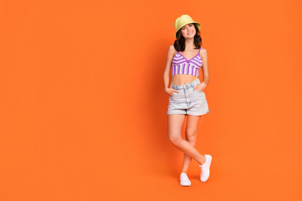 甜美的棕发女士穿着帽子、牛仔短裤和靴子，站立在橙色背景前的全身照片图片下载